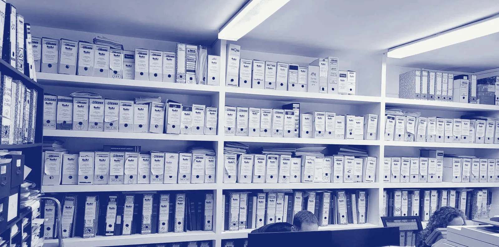 Pared del despacho con los archivadores de las comunidades, ordenados por número, y dos personas del equipo trabajando.